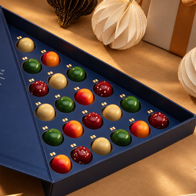 Open CACAO Christmas Chocolate Tree Praline Gift Box featuring 21 colourful chocolate bonbons in red, green, gold and orange, presented in navy blue packaging with festive paper ornaments against a warm, moody background.