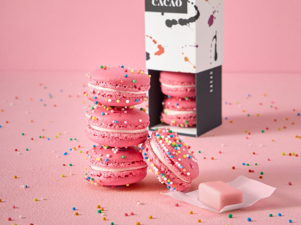 Stack of handcrafted Bubble Gum macarons with colourful sprinkles beside an open CACAO macaron gift box and a piece of pink bubble gum on a pastel pink background.