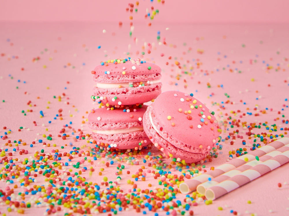 Handcrafted Bubble Gum macarons with colourful sprinkles surrounded by rainbow sprinkles and pink striped paper straws on a pastel pink background.