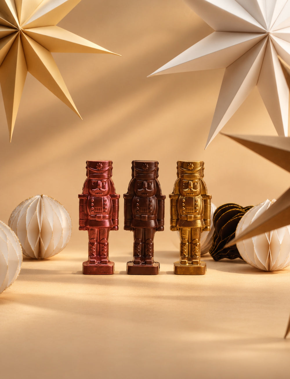 Three CACAO chocolate nutcrackers in pink, dark chocolate and gold, arranged with paper ornaments and large star decorations against a warm caramel-toned background.