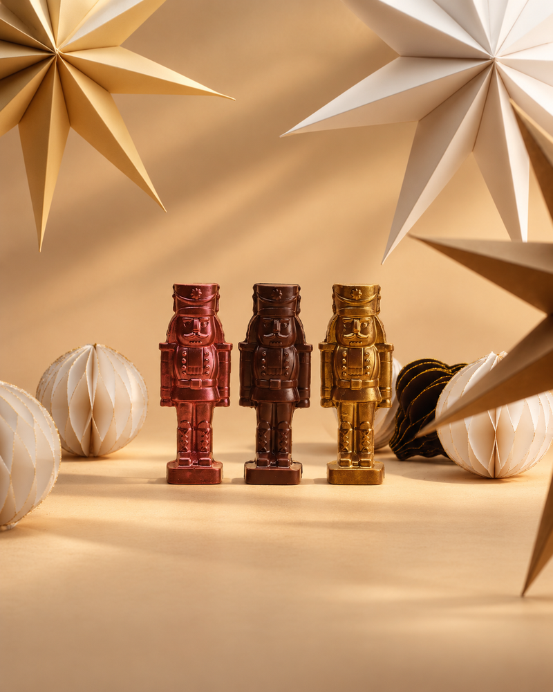 Three CACAO chocolate nutcrackers in metallic pink, dark chocolate and gold, surrounded by festive paper stars and ornaments against a softly lit, warm caramel background.