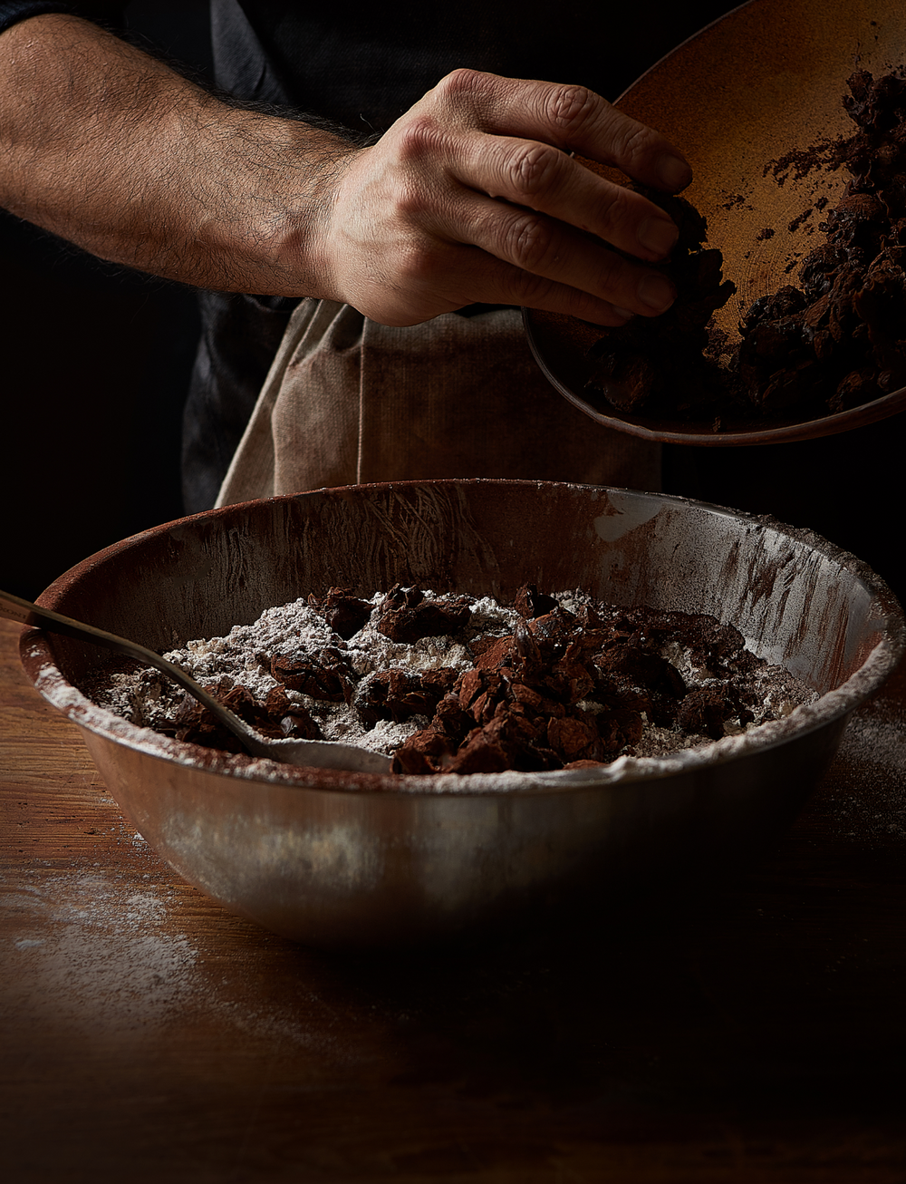 A close up of a chocolatier hand mixing dark cocoa powder into a bowl of chocolate in a moody setting